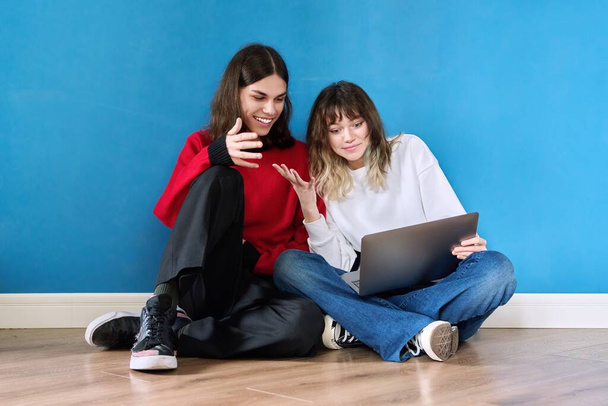 Couple of laughing cheerful teenage students using laptop sitting on the floor on blue background. E-education, online learning, youth, friendship, lifestyle, leisure, technology concept - Foto, Bild