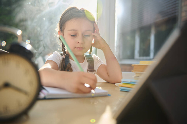View through a blurred alarm clock and tablet to a schoolgirl first grader learning the writing lesson, studying online in an online school. Homeschooling. Knowledge and education across the distance - Foto, imagen