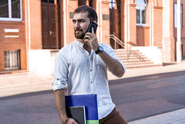 Уверенный красавчик. Молодой бизнесмен в белой рубашке разговаривает по телефону на улице. Самозанятый профессионал. Бизнес переговоры - Фото, изображение