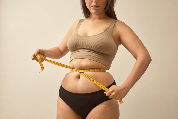 Woman with an excess of weight make waist with tape measure. Warm tones, studio shoot, natural normal female body. Body-shaming problem, - Foto, Imagem
