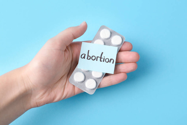 Woman holding paper note with word Abortion and pills on light blue background, top view - Foto, Imagem