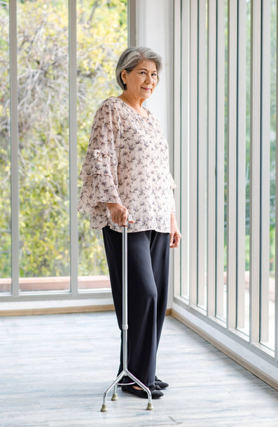 Full body shot Asian old senior happy healthy gray hair female retired pensioner in casual wear standing smiling in hospital hallway with glass windows using support assist walking cane stick walker. - Fotografie, Obrázek