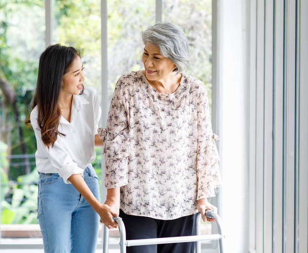 Asian young beautiful happy female granddaughter smiling helping cheerful old senior healthy gray hair retired pensioner grandmother walking using support assist four legged walker in living room. - Foto, Imagem