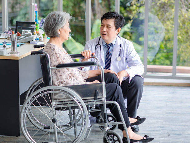 Asian happy male professional doctor in white lab coat with stethoscope visiting smiling talking monitoring old senior gray hair retired pensioner handicapped patient sitting on wheelchair in office. - Foto, Imagen