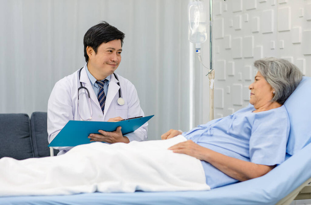 Профессиональный врач-азиат в белом халате и стетоскопе пишет заметки при посещении, наблюдая за пожилой бабушкой-пенсионеркой в больничной форме лежащей на кровати. - Фото, изображение