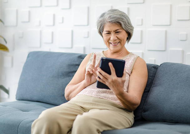 Asian cheerful old senior healthy gray hair female pensioner grandmother sitting smiling on cozy sofa in living room holding using touchscreen tablet computer surfing browsing application online. - Valokuva, kuva