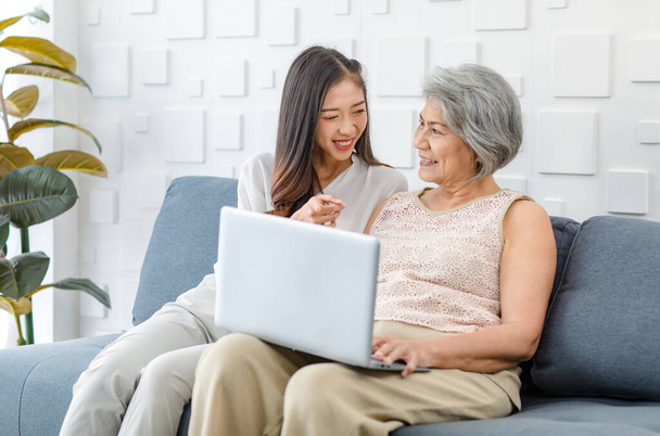 Ασιάτισσα χαρούμενη ηλικιωμένη υγιή γκρίζα μαλλιά γυναίκα συνταξιούχος μητέρα κάθεται χαμογελώντας με νεαρή κόρη σε άνετο καναπέ στο σαλόνι χρησιμοποιώντας φορητό υπολογιστή περιήγηση ψώνια online μαζί. - Φωτογραφία, εικόνα