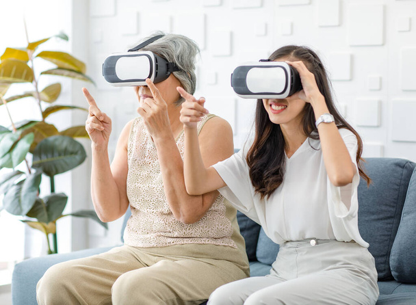 Asian young cheerful daughter wearing VR virtual reality goggles headset sitting smiling on cozy sofa in living room teaching old senior grey hair pensioner mother playing streaming 3d game online. - Foto, immagini