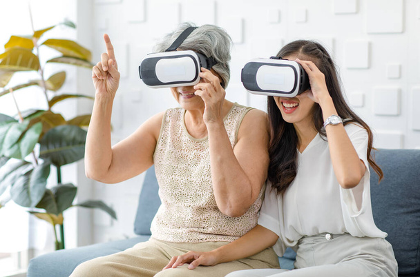 Азиатская юная жизнерадостная дочь в очках виртуальной реальности сидит улыбаясь на уютном диване в гостиной и учит пожилую мать пенсионера седой волосы играть в стриминг 3D игры онлайн. - Фото, изображение
