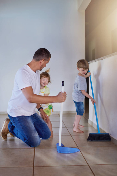 Η ομαδική δουλειά το κάνει όλο και πιο γρήγορα. Δύο μικρά αδέλφια βοηθούν τον πατέρα τους να σκουπίσει το πάτωμα στο σπίτι. - Φωτογραφία, εικόνα