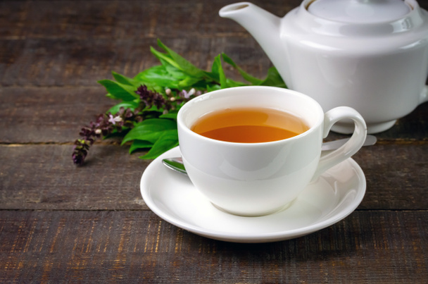Базиліка чаю в білій чашці кераміка з зеленим листом і білим чайником на сільському дерев'яному столі. Базилік це їжа і трава для здорового
. - Фото, зображення