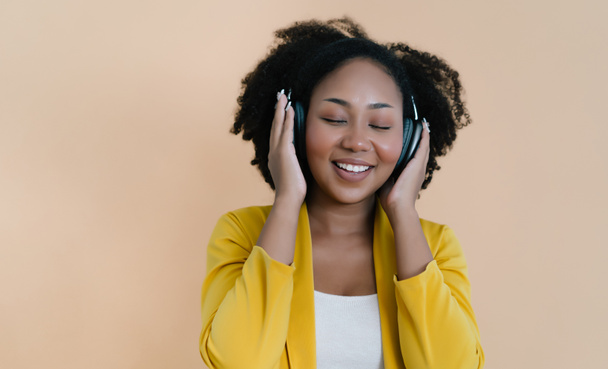 Fashionable Beautiful African American woman enjoy listening favorite music in headphones, closing her eyes from pleasure, music lover with smile on beige isolated studio background - 写真・画像