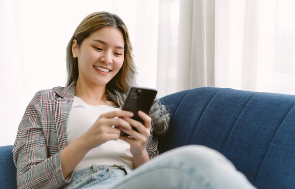 Улыбающаяся азиатская женщина использует смартфон для просмотра или просмотра социальных сетей дома. Свободная кореянка или японка, сидящая на удобном диване и расслабляющая. - Фото, изображение
