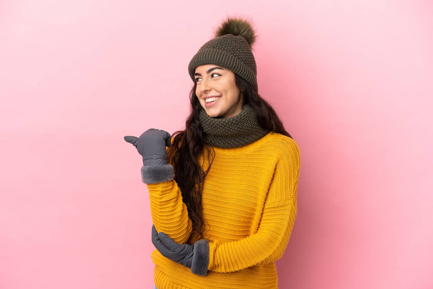 Young caucasian girl with winter hat isolated on purple background pointing to the side to present a product - Foto, Imagen