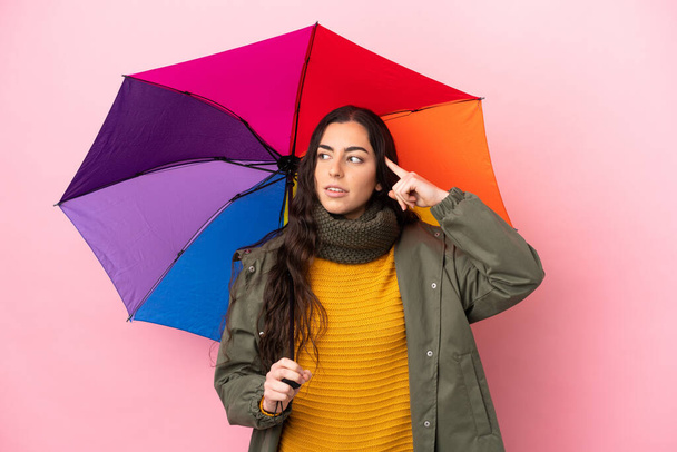 Молода жінка тримає парасольку ізольовано на рожевому тлі, маючи сумніви та мислення
 - Фото, зображення