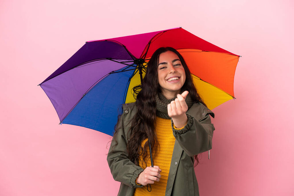 Młoda kobieta trzyma parasol odizolowany na różowym tle wykonując gesty pieniężne - Zdjęcie, obraz