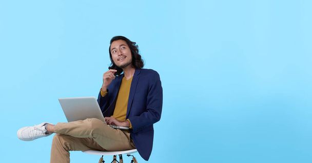 Young asian businessman thinking. While her using laptop sitting on  chair and looking isolate on blue background. - Foto, Imagem