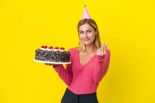 Блондинка Уругвая держит торт на день рождения изолирован на желтом фоне, зарабатывая деньги жест - Фото, изображение