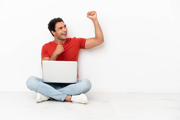 Caucasian handsome man with a laptop sitting on the floor celebrating a victory - 写真・画像