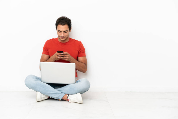 Белый красавчик с ноутбуком, сидящим на полу, отправляет сообщение с мобильным телефоном - Фото, изображение