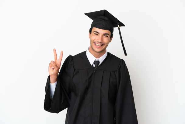 Jong universitair afgestudeerde over geïsoleerde witte achtergrond glimlachen en tonen overwinning teken - Foto, afbeelding