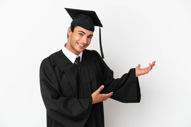 Молодой выпускник университета на изолированном белом фоне протягивает руки в сторону для приглашения приехать - Фото, изображение