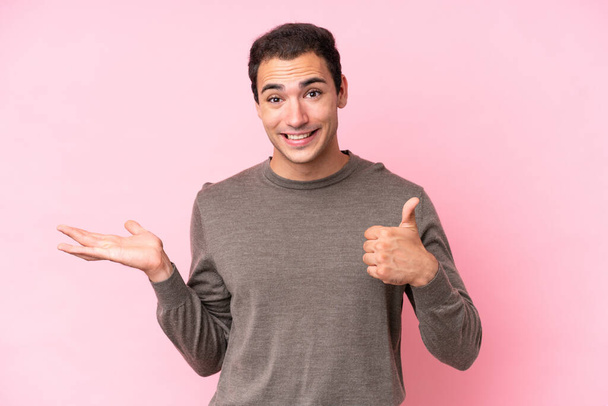 Young caucasian man isolated on pink background holding copyspace imaginary on the palm to insert an ad and with thumbs up - Foto, immagini