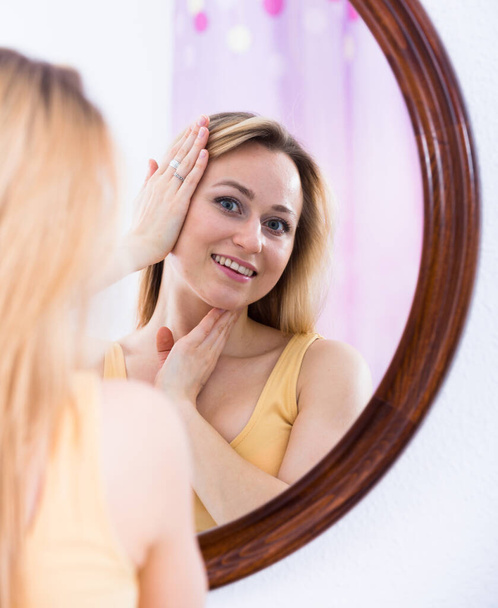Nuori blondi nainen tarkkaavaisesti katselee itseään peilistä
 - Valokuva, kuva