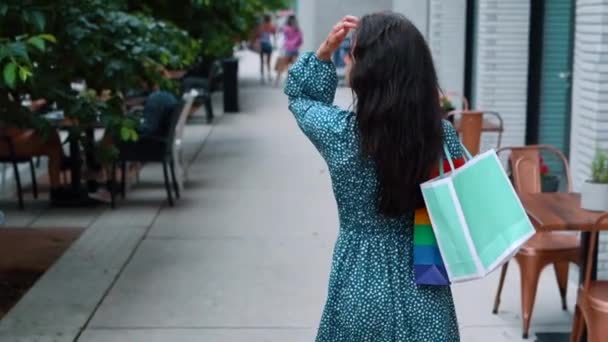 Rear of young pretty female in dress in polka dot walking on street with packets after shopping and turning her head to camera. Beautiful Caucasian woman with bags. Purchase concept. Slow motion - Imágenes, Vídeo