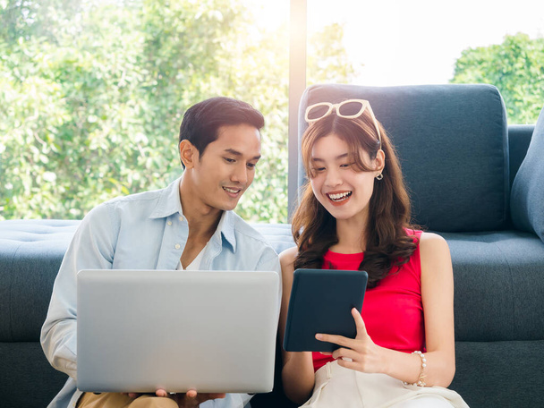 Happy Asian couple, young man and woman looking digital tablet screen while using laptop computer together for flight booking and trip information in living room, preparing to summer vacation travel. - Foto, imagen