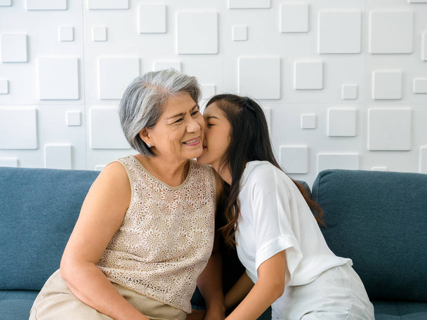 Блаженний азіатський старший, мати біле волосся обіймає свою прекрасну дочку, а вона цілує її щоку з любов'ю, турботою, комфортом і посмішкою, сидячи на дивані на білому фоні у вітальні вдома.. - Фото, зображення