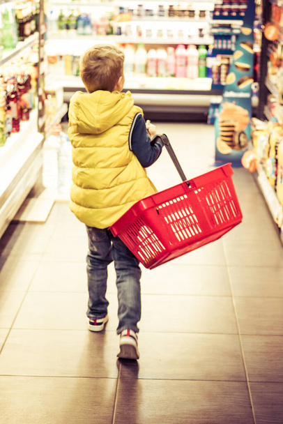 Rear view of boy with shopping basket walking through supermarket.  - Фото, зображення