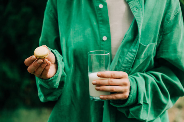 Γυναίκα σε πράσινο πουκάμισο κατέχουν μπισκότο μακαρόν και ένα ποτήρι γάλα - Φωτογραφία, εικόνα