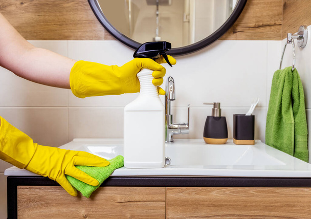 close-up of a female hand in a protective glove holding white bottle, cleaning agent mockup, space for logo and label. Bathroom background. The concept of hygiene, cleanliness and cleaning in house - Photo, image