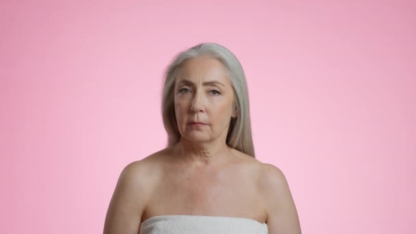 Aged beauty. Studio portrait of attractive grey haired senior woman wrapped in towel posing over pink background, slow motion, free space - Video