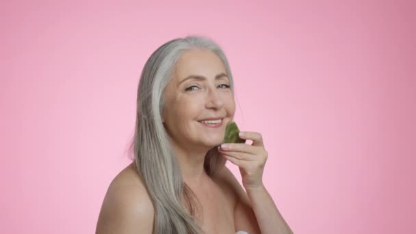 Beauty care procedures. Happy beautiful grey haired senior lady doing skin lifting massage with gouache stone, smiling to camera, pink studio background, slow motion - Séquence, vidéo