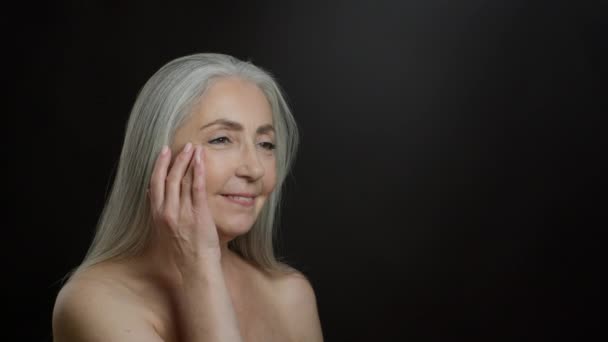 Γήρανση και ειδική κοσμετολογία. Studio πορτρέτο της όμορφης ηλικιωμένης γκριζομάλλης κυρίας τρίβει την κρέμα ανύψωσης του δέρματος στο πρόσωπό της, ποζάροντας με γυμνούς ώμους πάνω από μαύρο φόντο, αργή κίνηση, κενό χώρο - Πλάνα, βίντεο