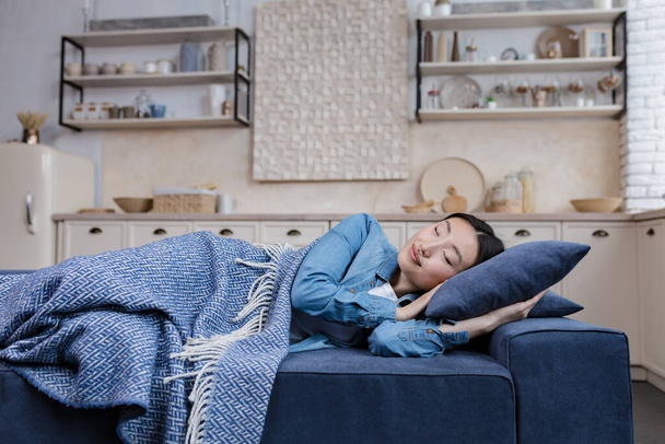 Young beautiful Asian woman resting and sleeping at home lying on sofa under blanket in living room, day nap - 写真・画像