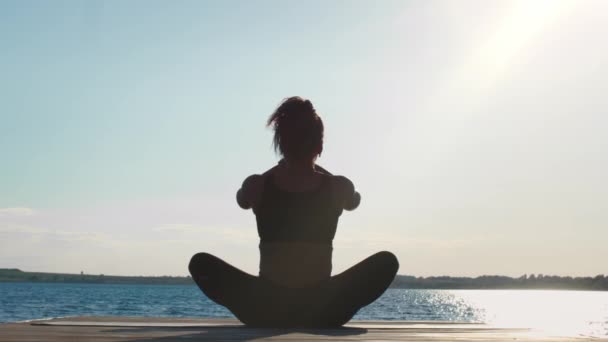 Единство с природой. Вид сзади на спокойную черную женщину, медитирующую на открытом воздухе, сидящую на деревянном пирсе возле большой воды утром, замедленное движение, свободное пространство - Кадры, видео