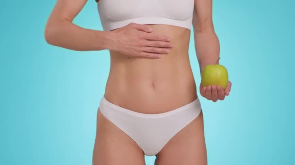 Balanced digestive process. Close up shot of slim unrecognizable woman in underwear stroking her fit belly, holding green organic apple, blue studio background, slow motion - Metraje, vídeo