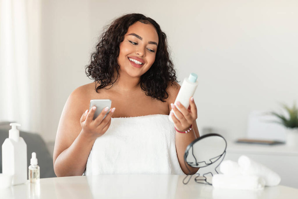 Happy black chubby woman in white towel reading recommendations on lotion bottle and using smartphone, sitting in bedroom interior and smiling - Fotó, kép