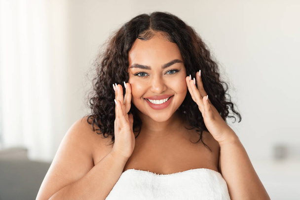 Face massage. African american plus size woman touching her smooth flawless skin on cheeks and smiling at camera, sitting in bedroom interior, wrapped in towel - Photo, Image