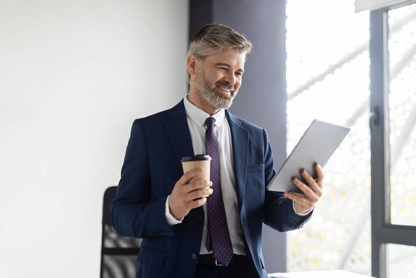 Portrait Of Smiling Middle Aged Businessman Drinking Coffee And Using Digital Tablet In Office, Happy Handsome Mature Man In Suit Relaxing At Workplace With Modern Gadget, Enjoying Work Break - Valokuva, kuva