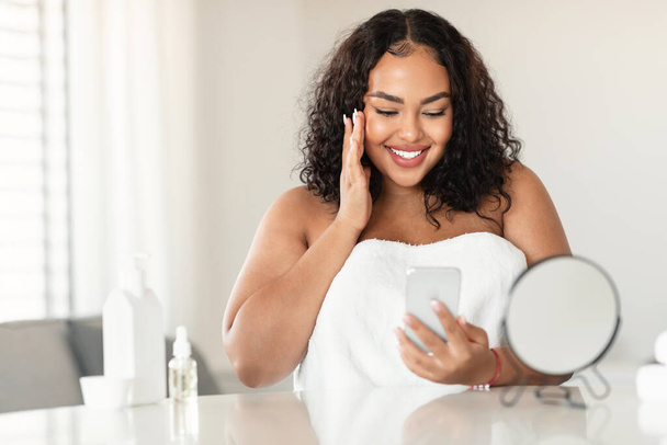 Ελκυστική μαύρη bodypositive γυναίκα σε λευκή πετσέτα κάθεται στην κρεβατοκάμαρα, αγγίζοντας το πρόσωπό της και χαμογελώντας, διαβάζοντας το blog ομορφιάς ή βλέποντας βίντεο σε απευθείας σύνδεση, χρησιμοποιώντας smartphone, ελεύθερο χώρο - Φωτογραφία, εικόνα