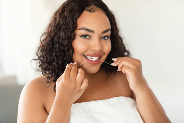 Young black chubby woman cleaning teeth with dental floss at home and looking at camera. Happy plus size lady performing everyday dental care. Oral hygiene, healthy lifestyle concept - Foto, afbeelding