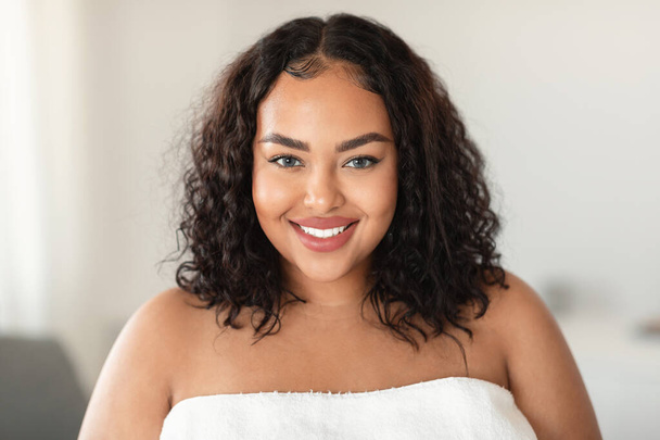 Beauty portrait of african american oversize lady standing wrapped in towel and smiling at camera, happy black woman with smooth skin posing after bath - Foto, afbeelding