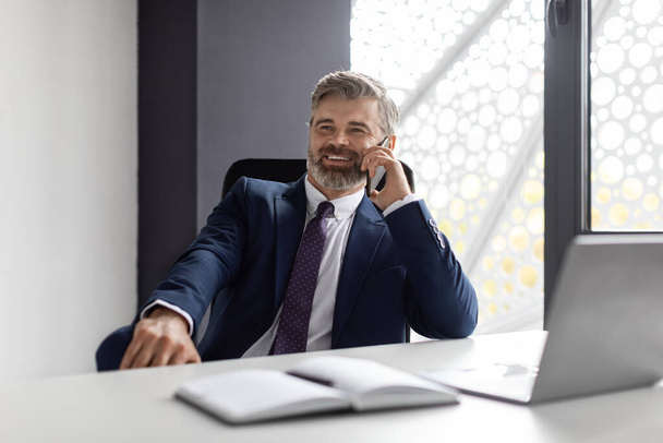 Phone Call. Smiling Mature Businessman In Suit Talking On Cellphone At Workplace, Handsome Relaxed Male Entrepreneur Sitting At Desk In Office, Enjoying Pleasant Mobile Conversation, Free Space - Zdjęcie, obraz