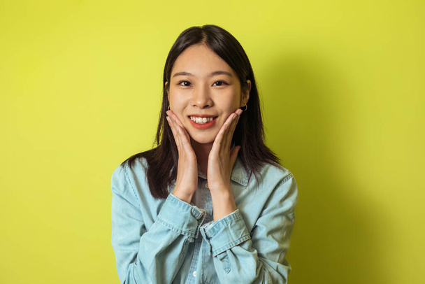 Portrait Of Cheerful Japanese Woman Touching Face Smiling Looking At Camera Stosing Over Yellow Studio Background. Женская красота и уход за кожей лица - Фото, изображение