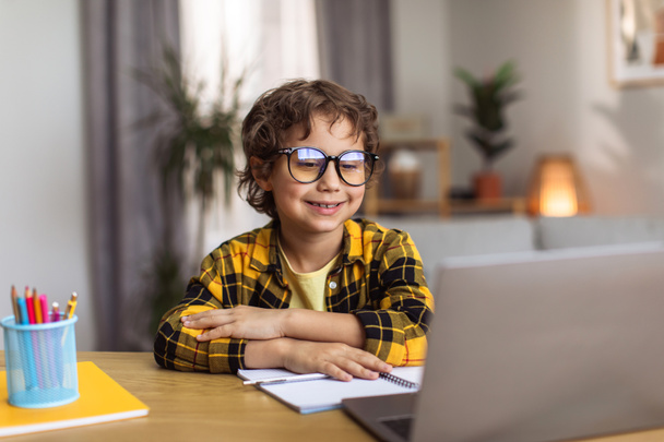 Happy learning. Clever little boy wearing eyeglasses watching lesson online on laptop, sitting at home workplace and smiling, free space - Fotó, kép