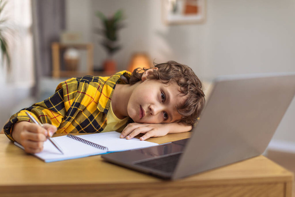 Βαρετός αναστατωμένος μικρό αγόρι που βρίσκεται στο χέρι και γράφοντας σε copybook, βλέποντας το μάθημα σε απευθείας σύνδεση στο laptop στο σπίτι, άδειο χώρο - Φωτογραφία, εικόνα